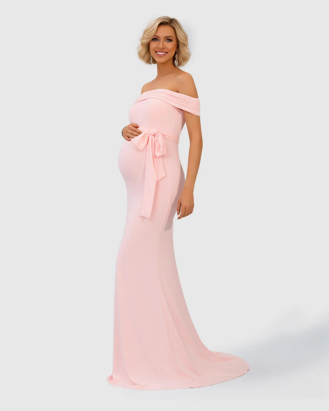 Blonde pregnant woman in a pink off-shoulder maternity gown with an adjustable tie-waist and flowing fishtail hem.