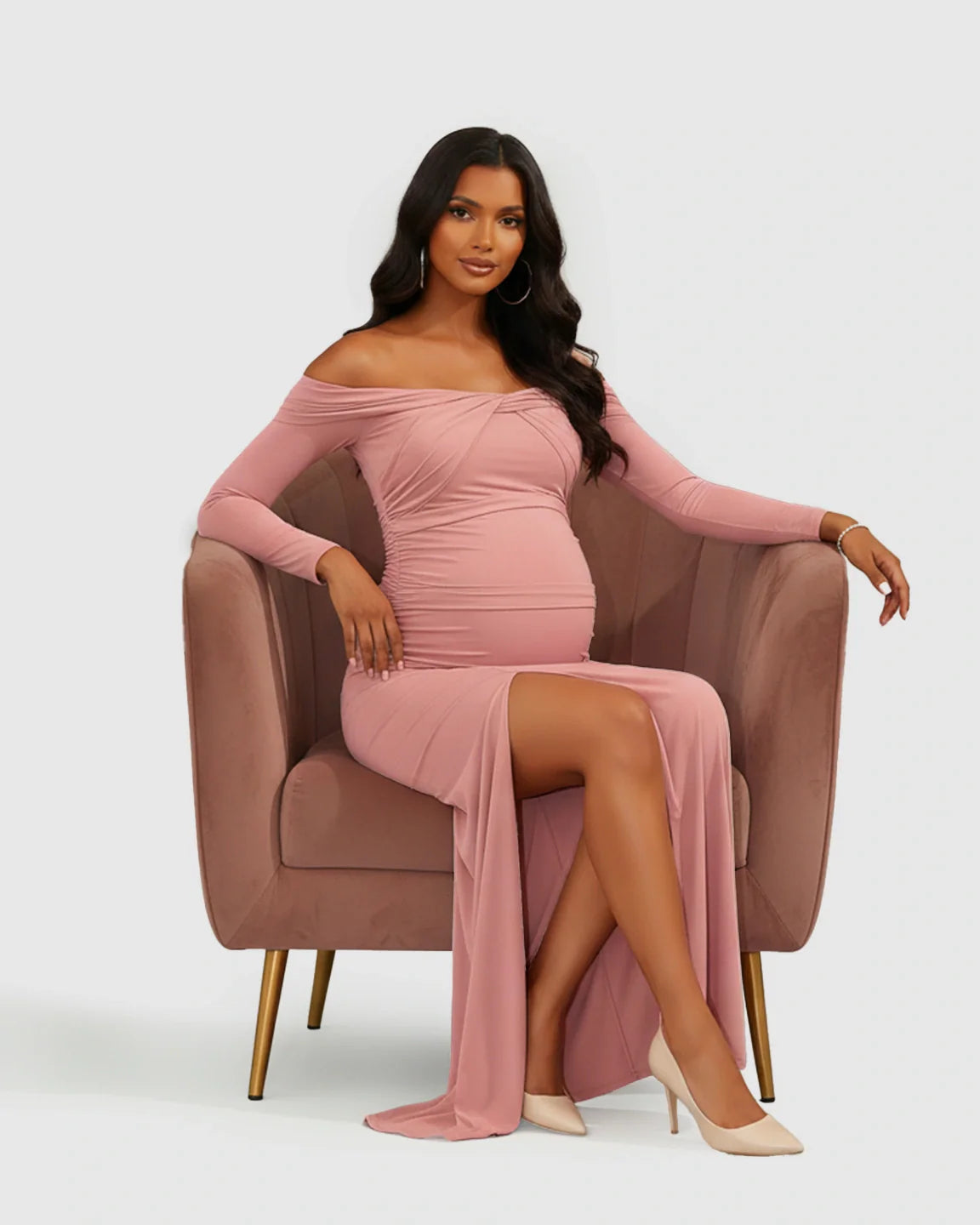 Maternity model in a pink ruched off-shoulder bodycon dress with a high slit, sitting in a velvet chair.